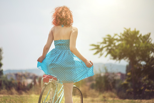 Rare view of curly red haired girl in retro styled blue dress. She is sitting on retro blue bike  in spectacular natural scenery, on warm summer day.