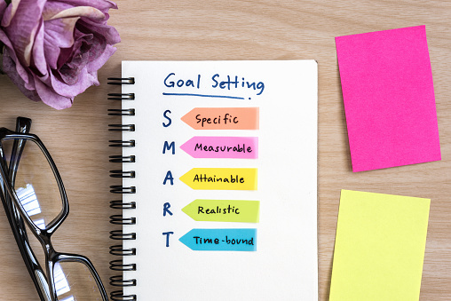 Hand writing definition for smart goal setting on notebook with eye glasses, purple rose and colorful sticky note on desk