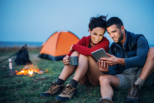 Smiling man and woman resting next to the campfire and using digital tablet