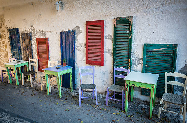 Green and white tables and white and purple seats near blue, green and red windows on a street in Crete island, Greece.