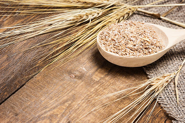 Wheat bran in wooden spoon with wheat ears Close-up of wooden spoon filled with wheat bran surrounded by wheat ears on burlap fabric and wooden planks. Dietary supplement to improve digestion. Source of dietary fiber. Shallow depth of field bran stock pictures, royalty-free photos & images