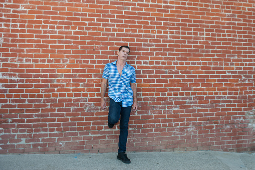 Male model standing with leg up on red brick wall.