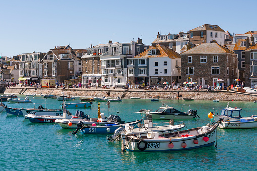 St Ives, England - July 3, 2016: St Ives harbour at high tide in Cornwall. Small boats are moored in the harbour and the historic buildings of St Ives can be seen lining the quayside where there are some pedestrians walking.