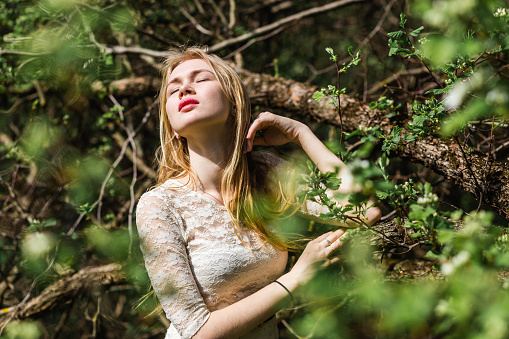 Portrait of a beautiful young blonde woman with long hair in a white dress on a background of blossom trees in spring, with closed eyes.