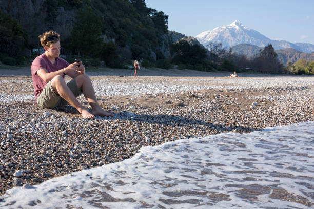 istock Young man sends text from beach, at sea edge 550207801