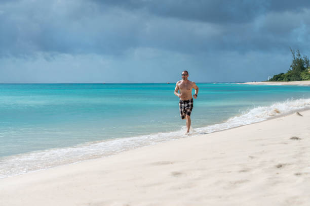 l'uomo corre lungo la spiaggia vuota sul bordo del surf - men beach running shirtless foto e immagini stock