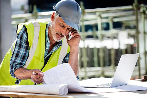 Male architect using mobile phone while reading documents at construction site