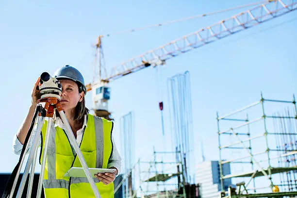 Female architect examining construction site through theodolite while holding digital tablet