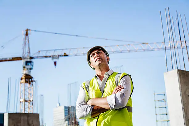 Ambitious male architect looking away while standing arms crossed at construction site against clear sky