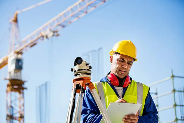 Male architect writing on clipboard by theodolite at construction site