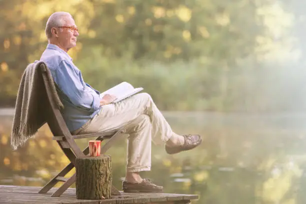 Photo of Older man reading book on dock at lake