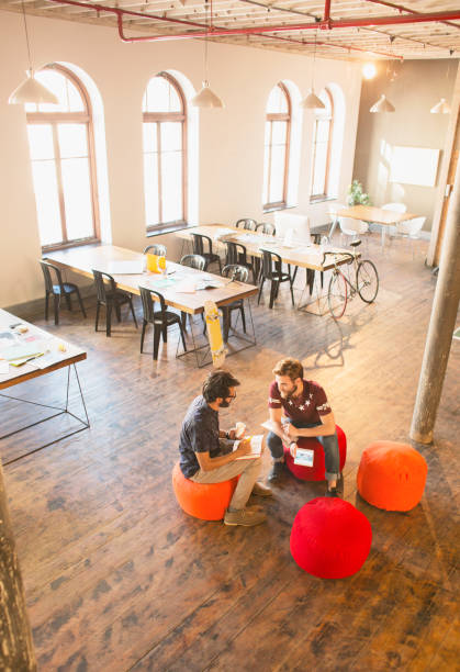 Comfy collaborative workspace. Open coworking space interior. Relaxing lounge zone with soft couch armchairs low table. Diverse people working on notebooks at individual desks with seats in background