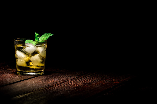 Drinking glass with ice cubes, mint and yellow liquid on the dark wooden surface.