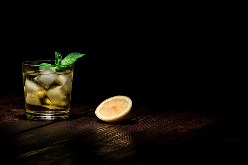 Drinking glass with ice cubes, mint, lemon and yellow liquid on the dark wooden surface.
