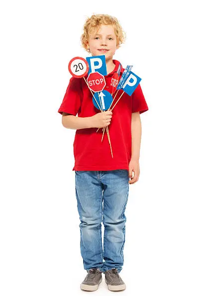 Full-length portrait of fair-haired schoolboy in red tee and denim, holding bunch of toy road signs, isolated on white