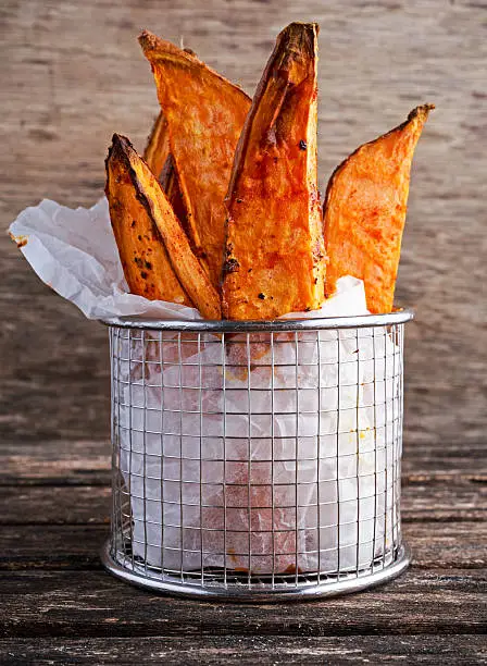 Freshly cooked sweet organic potato fries in paper wrap executed in a serving metal basket on old wooden table.