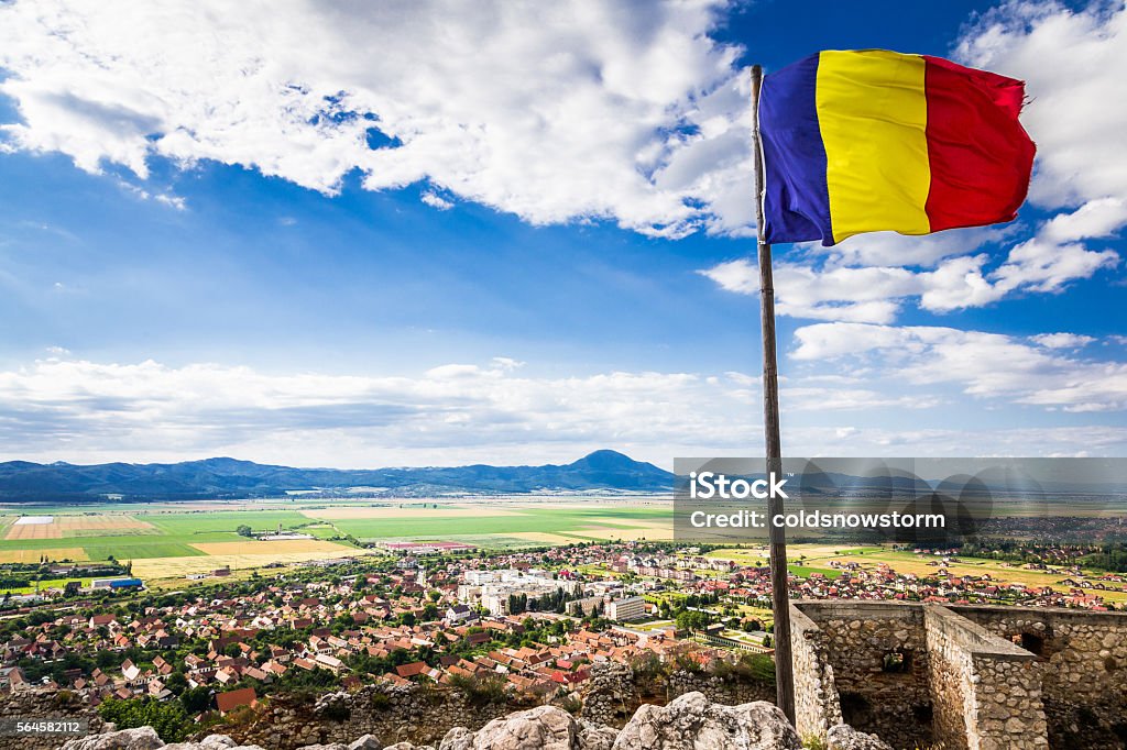 Romanian flag flying above the town of Rasnov, Transylvania, Romania Romanian flag flying above the town of Rasnov, Transylvania, Romania. The flag flies against a bright blue sky and cloudscape, with the rural town of Rasnov spread out below, with the mountains at the horizon beyond. The flag is located at the top of the famous citadel in Rasnov. Horizontal colour image. Romanian Flag Stock Photo