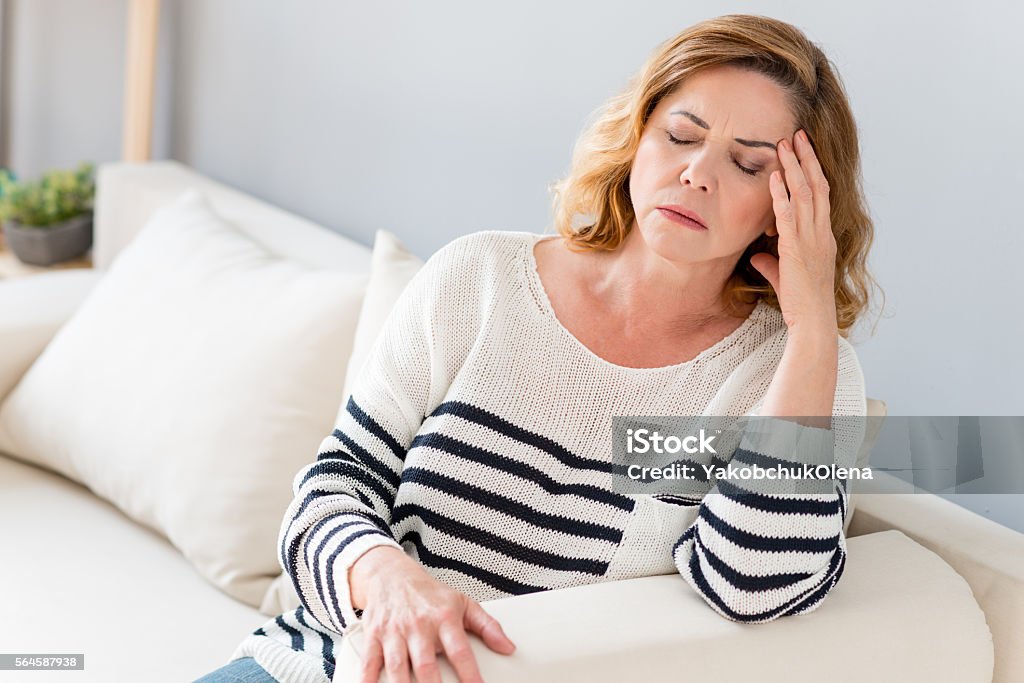 Senior lady feels pain in head Mature woman is suffering from headache. She is sitting on sofa and touching temple. Her eyes are closed with frustration Headache Stock Photo