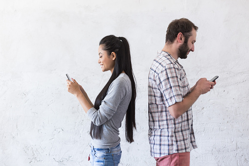 Cheerful young couple standing in empty room and using phone at new home, typing messages. Both wears casual clothes, standing back to back. Blank wall on background.