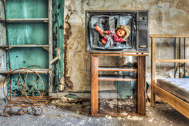 Smashed TV with doll inside screen in a dilapidated bedroom stock photo