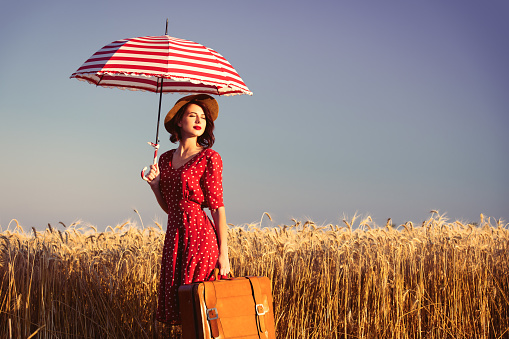 portrait of the beautiful young woman with red umbrella and brown suitcase standing in the field