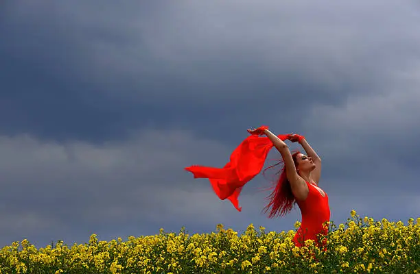  A young beautiful woman dressed in red, stands in a rape field and lets her scarf fly in the wind. The sky is stormy and dramatic. 