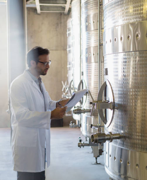 istock Vintner in lab coat with clipboard checking stainless steel vat in winery cellar 568518853