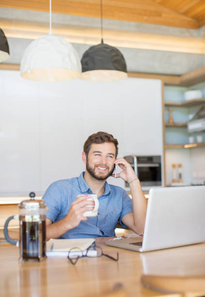 istock Smiling man drinking coffee and talking on cell phone at laptop 568519041