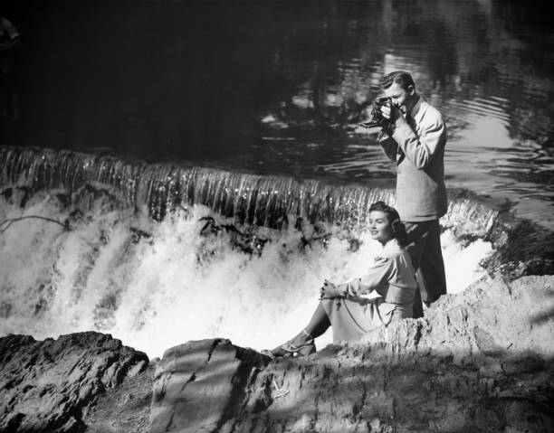 giovane coppia in cascata, uomo prendendo foto, (b & w - 1950s style couple old fashioned heterosexual couple foto e immagini stock