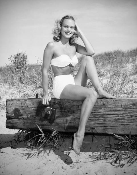 Woman wearing bikini sitting on log on beach, (B&W), (Portrait)  swimwear photos stock pictures, royalty-free photos & images