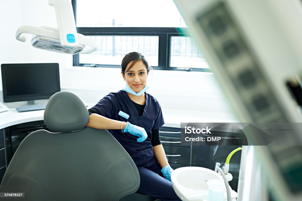 Dental assistant sitting on a dentist stool Portrait of attractive dental assistant sitting on a dentist stool at clinic looking at camera Dentist Stock Photo