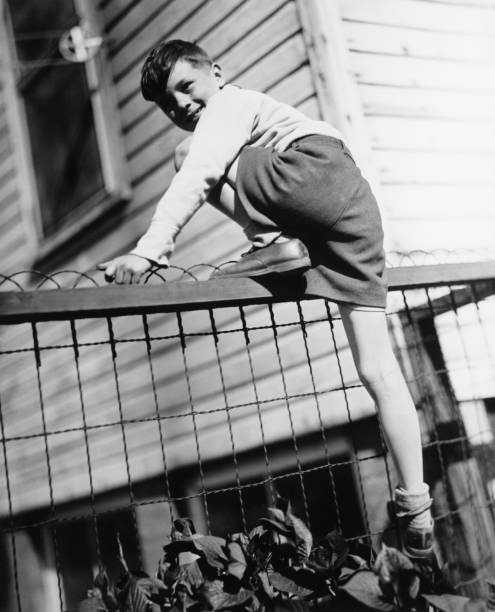 Boy (8-9) climbing on enclosure in front of house, (B&W)  clambering photos stock pictures, royalty-free photos & images