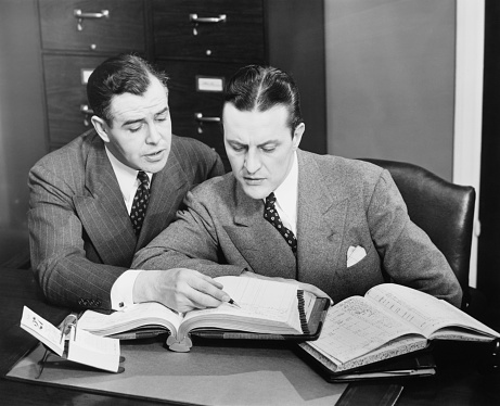 Two handsome young businessmen in classic suits are holding paper and examining document, standing outside the office building