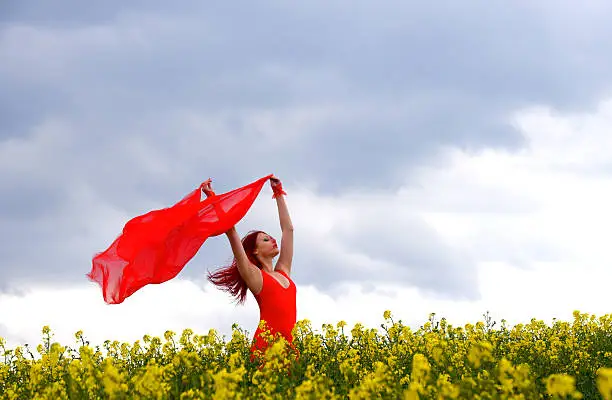  A young beautiful woman dressed in red stands in a rape field playing with her red scarf in the wind. She has her eyes closed. 
