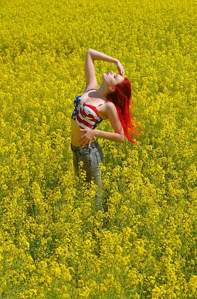 A beautiful young girl stands alone in a rape field in full bloom.  She wears a short stars and stripes top