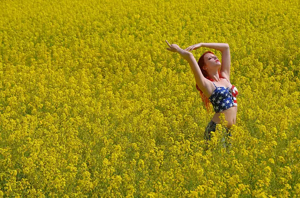 A beautiful young girl stands alone in a rape field in full bloom.  She wears a short stars and stripes top