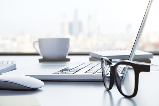 Closeup of white desktop with notebook, glasses, coffee cup, notepads and other items on blurry city background