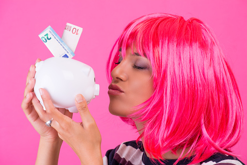 Young woman with pink wig kissing piggy bank on the pink background