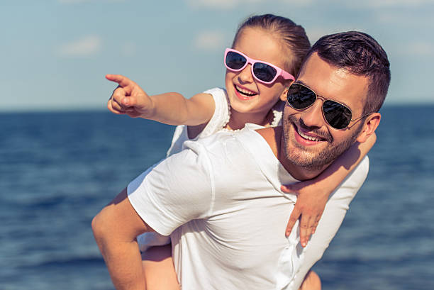 Young dad and his little daughter in sun glasses are looking away and smiling. Girl is sitting on her father's shoulders and pointing, sunny sea in the background