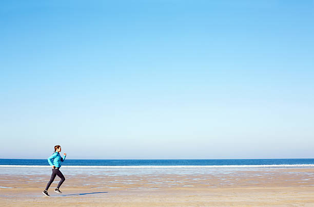 istock Fit woman jogging on beach against clear blue sky 576575382