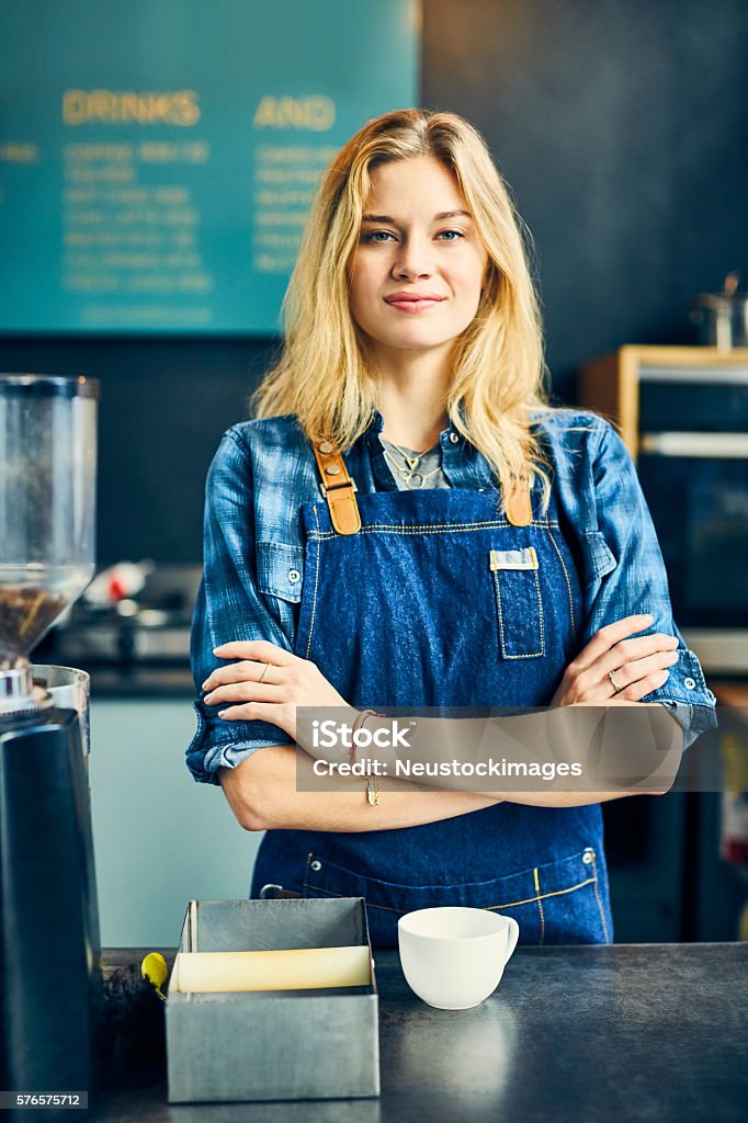 Attractive barista and owner standing arms crossed in cafe Portrait of attractive barista and owner with arms crossed at counter. Young female is smiling in coffee shop. She is wearing apron while standing in cafe. Blond Hair Stock Photo