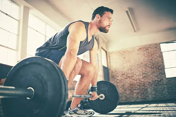 Full length of confident man dead lifting barbell in gym. Determined fit male is looking away while exercising. He is in sports wear.