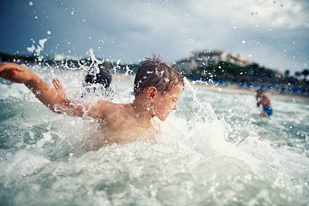 Little boy having fun splashing in sea waves. His brother and sister visible in background. Overcast day of summer vacations. 