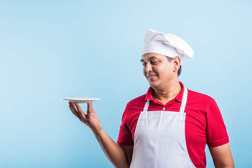 Portrait of handsome Indian male chef in uniform presenting empty plate and smiling, standing on plain blue background with copy space.