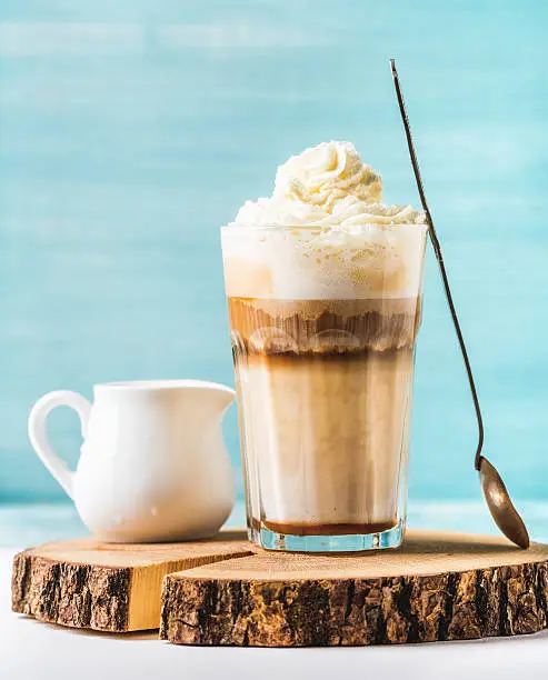 Latte macchiato with whipped cream, serving silver spoon and white pitcher on wooden round board over blue painted wall background, selective focus, vertical composition