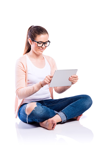 Teenage girl in white t-shirt, pink cardigan and jeans, sitting on the floor, holding tablet, writing on it, young beautiful woman, studio shot on white background, isolated