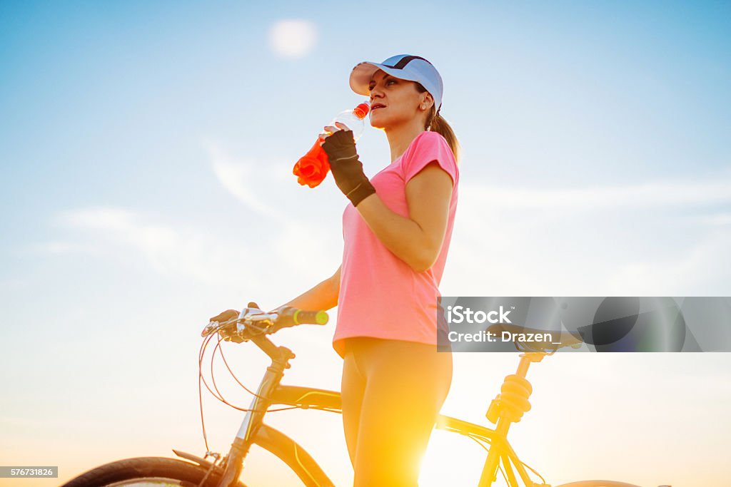 Young woman and recreation in summer afternoon with mountain bicycle Portrait of beautiful muscular woman holding grips of bike steering bar on sunny summer day in sunset, after exhausting ride and drinking energy drink. Woman is wearing cap and dressed in sports clothing. Location: Novi Sad, Serbia, Europe Drinking Stock Photo