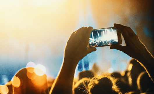 Somebody from the crowd is taping concert performance with a smartphone. Silhouette of hands holding the phone in focus with blurry stage and crowd in the background. Blue and orange toned image.