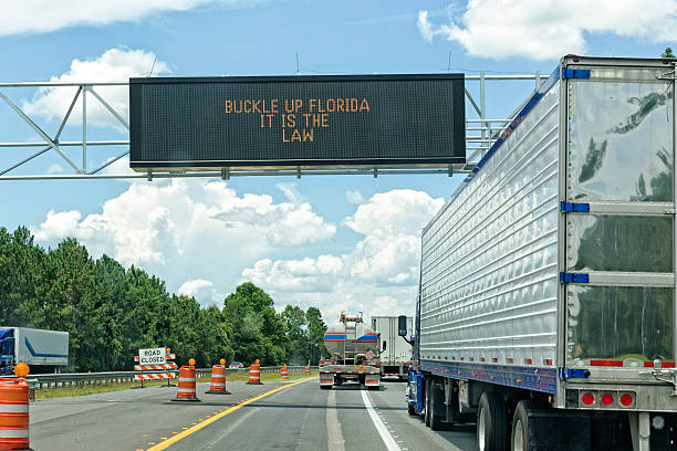 Signage on Highway 75 heading north telling drivers and passengers to buckle up in Florida. It is the law. Plenty of semi-trucks on a highway under construction. Taken from the inside of a car.