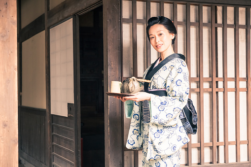 Retro revival image of a geisha walking in front of her house.  She's wearing a traditional kimono and brings a tea tray with tea mugs and teapot. Kyoto - Japan